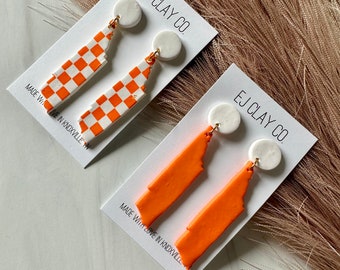 May include: Two pairs of orange and white Tennessee state-shaped earrings. The first pair is a checkerboard pattern and the second pair is solid orange. Both pairs have white round clay accents. The cards have the text "EJ CLAY CO. MADE WITH LOVE IN KNOXVILLE."
