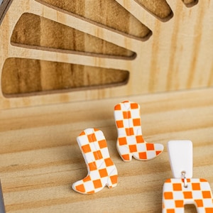 K&ouml;nnte beinhalten: Ein Paar orange-wei&szlig; karierte Cowboy-Stiefel-Ohrringe und ein passendes Paar rechteckiger Ohrringe. Die Ohrringe werden auf einer Holzoberfl&auml;che mit einem dekorativen Holz-Hintergrund pr&auml;sentiert.