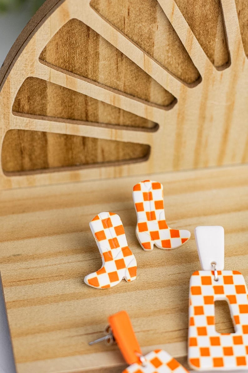 K&ouml;nnte beinhalten: Ein Paar orange-wei&szlig; karierte Cowboy-Stiefel-Ohrringe und ein passendes Paar rechteckiger Ohrringe. Die Ohrringe werden auf einer Holzoberfl&auml;che mit einem dekorativen Holz-Hintergrund pr&auml;sentiert.