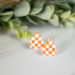 K&ouml;nnte beinhalten: Zwei kleine, k&uuml;rbisf&ouml;rmige Ohrringe mit einem wei&szlig;-orangenen Karomuster. Die Ohrringe sind auf einer wei&szlig;en Oberfl&auml;che platziert, mit gr&uuml;nem Laub im Hintergrund. Ein herbstliches Accessoire.