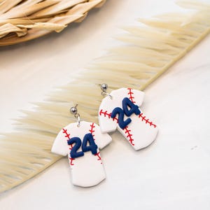 May include: A pair of white baseball jersey earrings with red stitching and a blue number 24. The earrings are hanging from silver hooks.