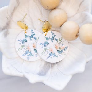 Pode incluir: Um par de brincos redondos brancos com uma cruz dourada e um design floral. Os brincos estão em um prato branco em forma de flor.