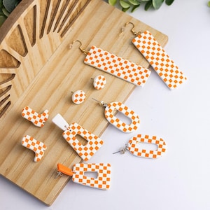 K&ouml;nnte beinhalten: Eine Sammlung handgefertigter Ohrringe in verschiedenen Formen, darunter Stecker, Rechtecke und Ovale. Die Ohrringe weisen ein wei&szlig;-oranges Karomuster auf. Sie werden auf einer Holzoberfl&auml;che mit einem dekorativen Holzakzent pr&auml;sentiert.