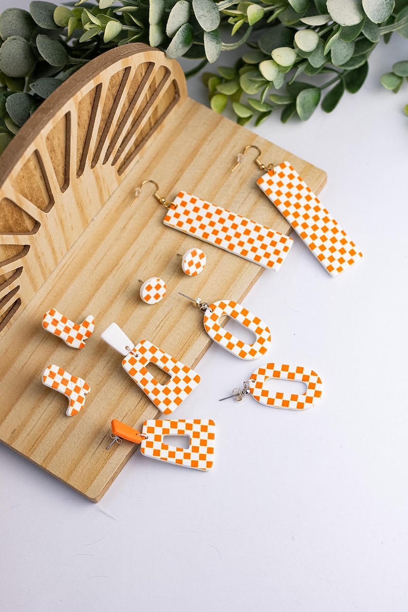 K&ouml;nnte beinhalten: Eine Sammlung handgefertigter Ohrringe in verschiedenen Formen, darunter Stecker, Rechtecke und Ovale. Die Ohrringe weisen ein wei&szlig;-oranges Karomuster auf. Sie werden auf einer Holzoberfl&auml;che mit einem dekorativen Holzakzent pr&auml;sentiert.