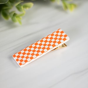 K&ouml;nnte beinhalten: Eine rechteckige Haarspange mit einem goldfarbenen Metallverschluss. Die Spange hat eine wei&szlig;e Basis mit einem sich wiederholenden orange-wei&szlig;en Schachbrettmuster. Die Spange ist abgewinkelt, und der Hintergrund ist unscharf.