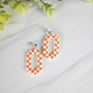 K&ouml;nnte beinhalten: Ein Paar orange-wei&szlig; karierte ovale Ohrringe. Die Ohrringe haben einen silbernen Stecker und eine hohle Mitte. Die Ohrringe sind vor einem wei&szlig;-grauen Marmorhintergrund mit gr&uuml;nem Laub in der oberen linken Ecke platziert.