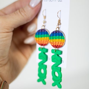 Puede incluir: Pendientes hechos a mano con un diseño de esfera arcoíris y la palabra "KNOX" en verde brillante. Los pendientes tienen ganchos dorados y se muestran sobre un fondo blanco.