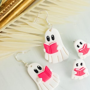 Op de afbeelding: Witte spookoorbellen met zwarte ogen en een roze boek. De oorbellen zijn gemaakt van lichtgewicht materiaal en hebben zilveren haken. De oorbellen zijn een leuk, spookachtig accessoire.