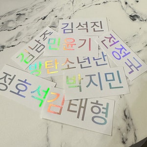 Può includere: Una collezione di adesivi olografici iridescenti con parole coreane, tra cui "Kim Seon Jin", "Jeong Ha", "Kim Yu Gi", "Baek Ji Min", "Yang Tae Hyung", "Gilman", "Batan Son Dan" e "Jeong Gyo".