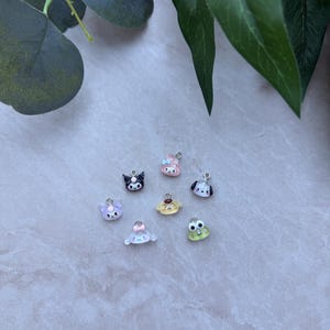 May include: A collection of small, colorful charms featuring various cartoon characters. The charms include a black cat, a pink bunny, a dog, a frog, and other cute designs. Each charm has a silver loop for attaching to jewelry or other crafts.
