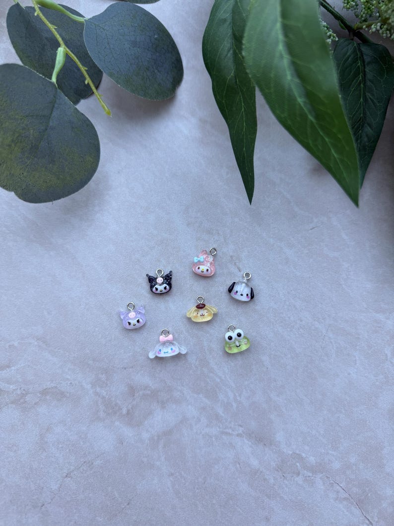 May include: A collection of small, colorful charms featuring various cartoon characters. The charms include a black cat, a pink bunny, a dog, a frog, and other cute designs. Each charm has a silver loop for attaching to jewelry or other crafts.