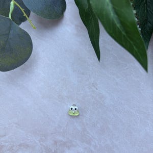 May include: A small, light green frog charm with black eyes and a silver-colored loop. The charm is set against a light gray background, with green leaves in the upper corners.