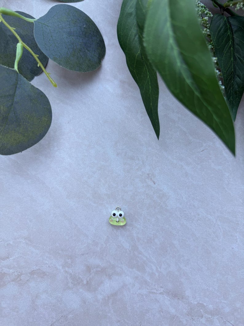 May include: A small, light green frog charm with black eyes and a silver-colored loop. The charm is set against a light gray background, with green leaves in the upper corners.