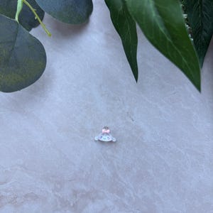 May include: A small, white charm with a light blue face and a pink bow. The charm has a cartoon-like design and is set against a light gray marble background, with green leaves in the upper corners.