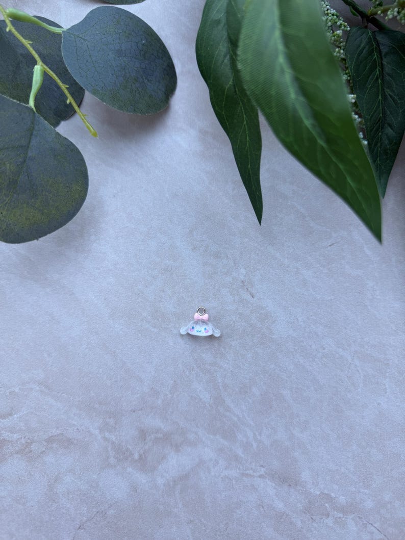 May include: A small, white charm with a light blue face and a pink bow. The charm has a cartoon-like design and is set against a light gray marble background, with green leaves in the upper corners.
