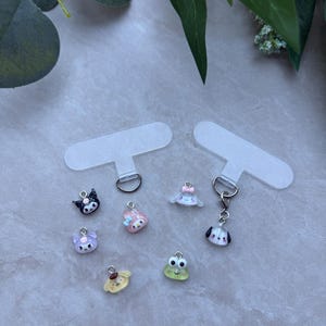 May include: A collection of small, colorful charms featuring various cartoon characters, including a black cat, a pink bunny, and a green frog. The charms are attached to clear phone grip holders, perfect for adding a touch of personality to your phone.
