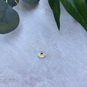 May include: A small, yellow charm with a red accent and silver-colored loop. The charm has a cartoon-like design. The background is a light gray surface with green leaves.
