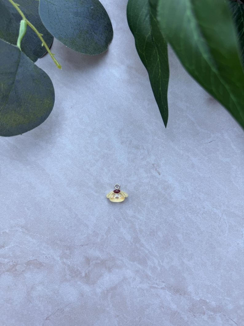 May include: A small, yellow charm with a red accent and silver-colored loop. The charm has a cartoon-like design. The background is a light gray surface with green leaves.