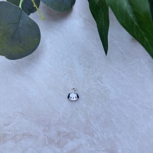 May include: A small, white and black dog charm with a silver-colored loop for attaching to a necklace or bracelet. The charm has a cute, cartoon-like design with a red nose and black ears. The background is a light, textured surface with green leaves.