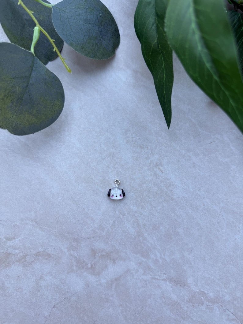 May include: A small, white and black dog charm with a silver-colored loop for attaching to a necklace or bracelet. The charm has a cute, cartoon-like design with a red nose and black ears. The background is a light, textured surface with green leaves.