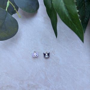 May include: Two small, cat-shaped charms. One is lavender with white accents, the other is black with white accents. Both have silver-colored loops at the top for attaching to jewelry. The charms are on a light gray surface, with green leaves in the background.
