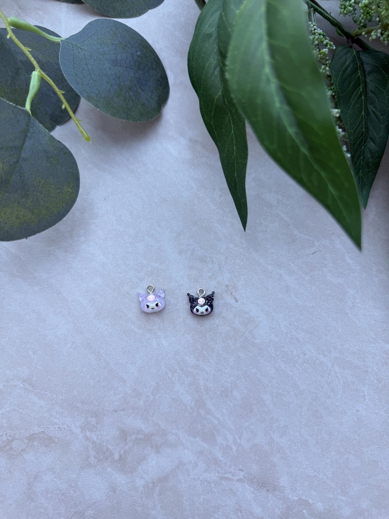 May include: Two small, cat-shaped charms. One is lavender with white accents, the other is black with white accents. Both have silver-colored loops at the top for attaching to jewelry. The charms are on a light gray surface, with green leaves in the background.