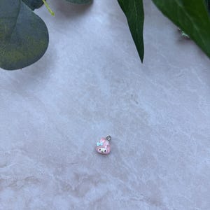 May include: A small, pink charm with a cartoon bunny design. The charm has a silver-colored loop at the top for attaching to a necklace or bracelet. The bunny has a blue bow and a smiling face.