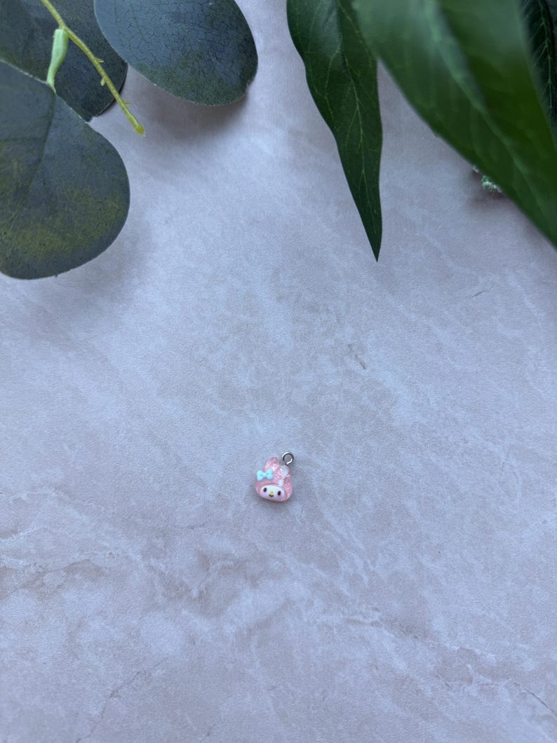 May include: A small, pink charm with a cartoon bunny design. The charm has a silver-colored loop at the top for attaching to a necklace or bracelet. The bunny has a blue bow and a smiling face.