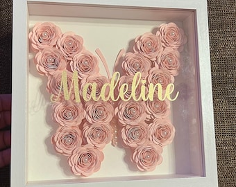 May include: A white frame with a pink paper butterfly made of layered circles. The butterfly has pearl accents and the name "Madeline" is written in gold lettering.