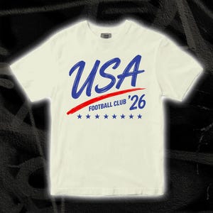Puede incluir: Camiseta color crema con la palabra "USA" en azul, una raya roja y el texto "FOOTBALL CLUB '26" en azul. Siete estrellas azules debajo. Una camiseta informal para los aficionados al f&uacute;tbol.