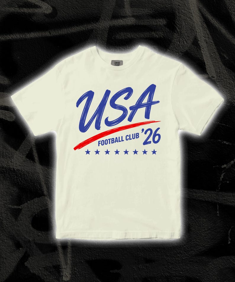 Puede incluir: Camiseta color crema con la palabra "USA" en azul, una raya roja y el texto "FOOTBALL CLUB '26" en azul. Siete estrellas azules debajo. Una camiseta informal para los aficionados al f&uacute;tbol.