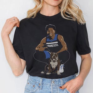 May include: Black t-shirt with a graphic of a basketball player wearing a blue and white jersey with the word "MINNESOTA" on it. The player is holding a brown and white bulldog.