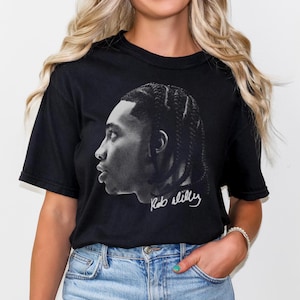 Puede incluir: Camiseta negra con una imagen en blanco y negro de un rostro de hombre de perfil. El hombre tiene el pelo trenzado y el texto "Rob Sills" está escrito debajo de la imagen.