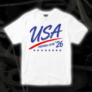 Puede incluir: Camiseta blanca con la palabra "USA" en azul manuscrito, una raya roja y el texto "FOOTBALL CLUB '26". Debajo hay siete estrellas azules. La camiseta tiene cuello redondo.
