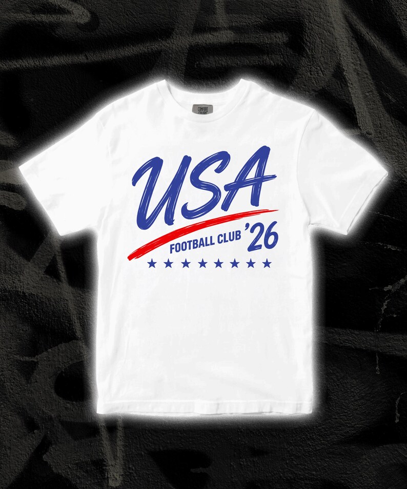 Puede incluir: Camiseta blanca con la palabra "USA" en azul manuscrito, una raya roja y el texto "FOOTBALL CLUB '26". Debajo hay siete estrellas azules. La camiseta tiene cuello redondo.