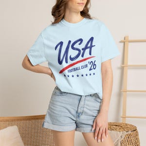 Puede incluir: Camiseta azul claro con el texto "USA Football Club '26" en azul marino y rojo. La camiseta tiene cuello redondo y mangas cortas. El dise&ntilde;o incluye una raya roja y estrellas azules.