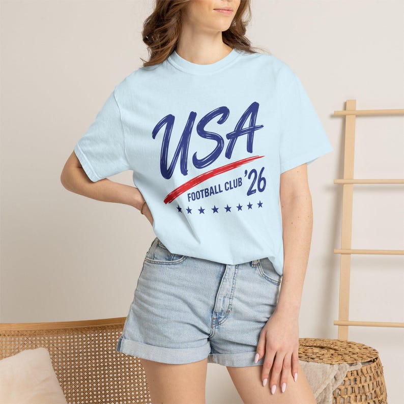 Puede incluir: Camiseta azul claro con el texto "USA Football Club '26" en azul marino y rojo. La camiseta tiene cuello redondo y mangas cortas. El dise&ntilde;o incluye una raya roja y estrellas azules.