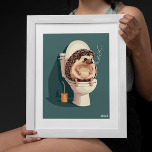 Könnte beinhalten: Ein weiß gerahmter Druck eines Comic-Igels, der auf einer Toilette sitzt und eine Zigarette im Maul hält. Der Hintergrund ist blaugrün und der Text "able6" befindet sich in der rechten unteren Ecke.