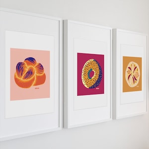 May include: Three framed wall art prints featuring illustrations of food items. The first shows a braided bread on a pink background, the second a speckled bagel on a magenta background, and the third a sliced onion on an orange background. Each print includes the text "abieé".