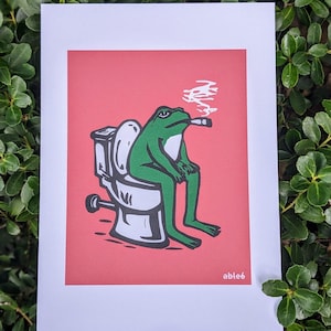 May include: A framed art print featuring a green frog smoking a cigar while sitting on a toilet. The frog is depicted in a relaxed pose against a red background. The print has the text "able6" in the bottom right corner.
