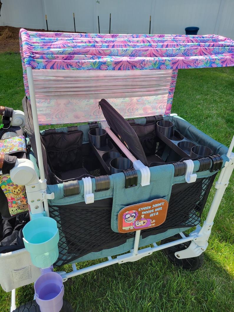 May include: A teal wagon with a black mesh storage area and a pink and white floral canopy. The wagon has multiple cup holders and a patch that says "Every Janet needs her ETA".