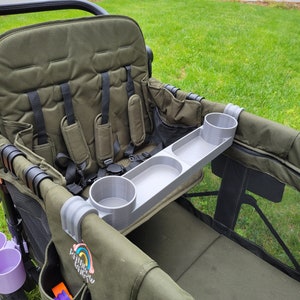 May include: A gray plastic tray with two cup holders attached to a green wagon. The tray is designed to fit over the side of the wagon and has a raised lip to prevent items from falling off. The wagon has a black frame and a green canvas seat.