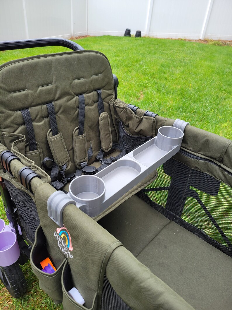 May include: A gray plastic tray with two cup holders attached to a green wagon. The tray is designed to fit over the side of the wagon and has a raised lip to prevent items from falling off. The wagon has a black frame and a green canvas seat.