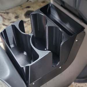 May include: Black plastic car console organizer with two cup holders and a storage compartment. The organizer is designed to fit in a vehicle's center console area. The item is attached with screws.