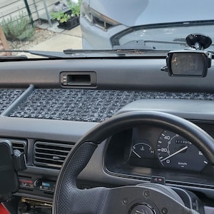 May include: Interior view of a car dashboard. The dashboard is gray with a textured, patterned insert. A black steering wheel, speedometer, and air vents are visible. A phone mount and a small electronic device are attached.