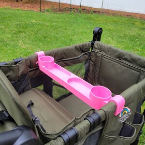 May include: Pink cup holder accessory for a wagon with two cup holders and a tray. The cup holder is attached to the side of the wagon with a black strap. The wagon is green and has the text "Rainbow Baby" on the side.