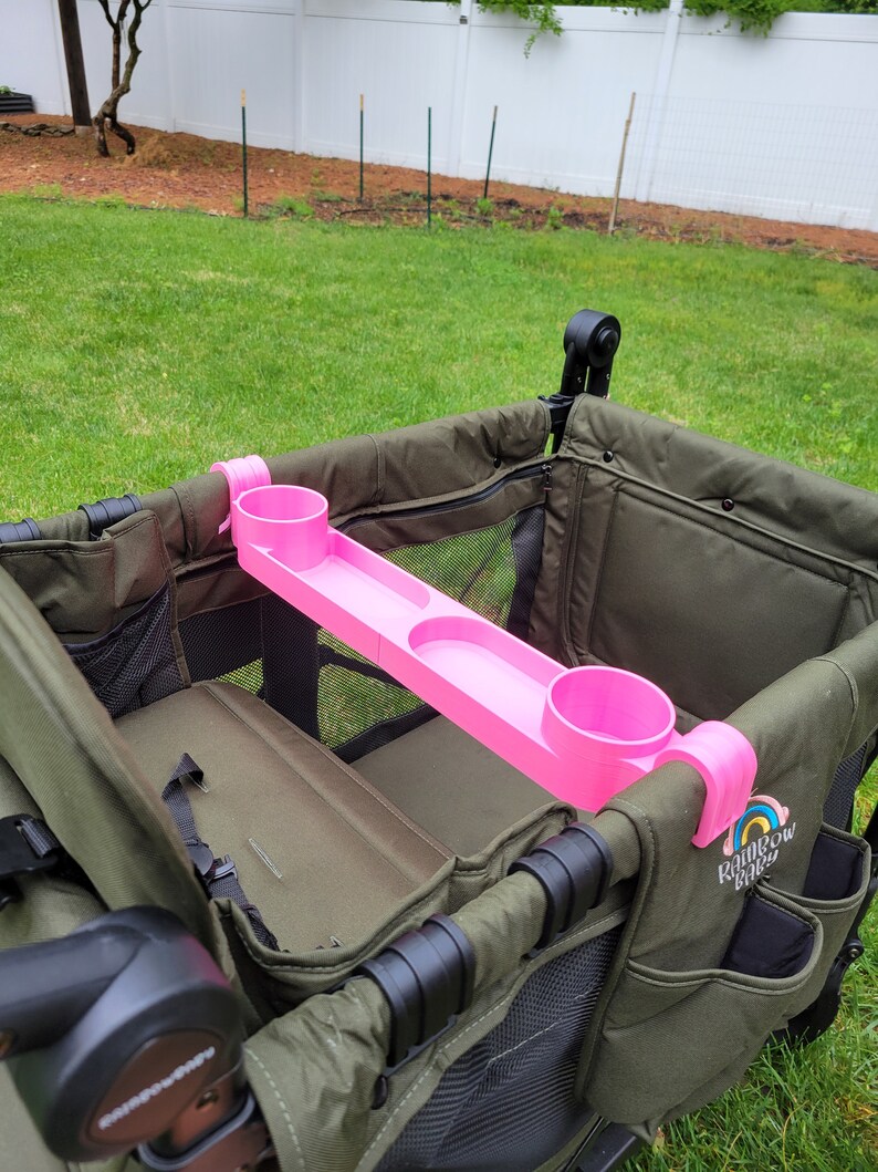 May include: Pink cup holder accessory for a wagon with two cup holders and a tray. The cup holder is attached to the side of the wagon with a black strap. The wagon is green and has the text "Rainbow Baby" on the side.