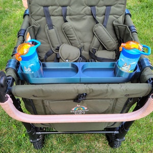 May include: A green wagon with two seats and a blue tray with two cup holders. The wagon has a pink handle and the words "Rainbow Baby" are printed on the side. Two blue water bottles with orange lids are in the cup holders.