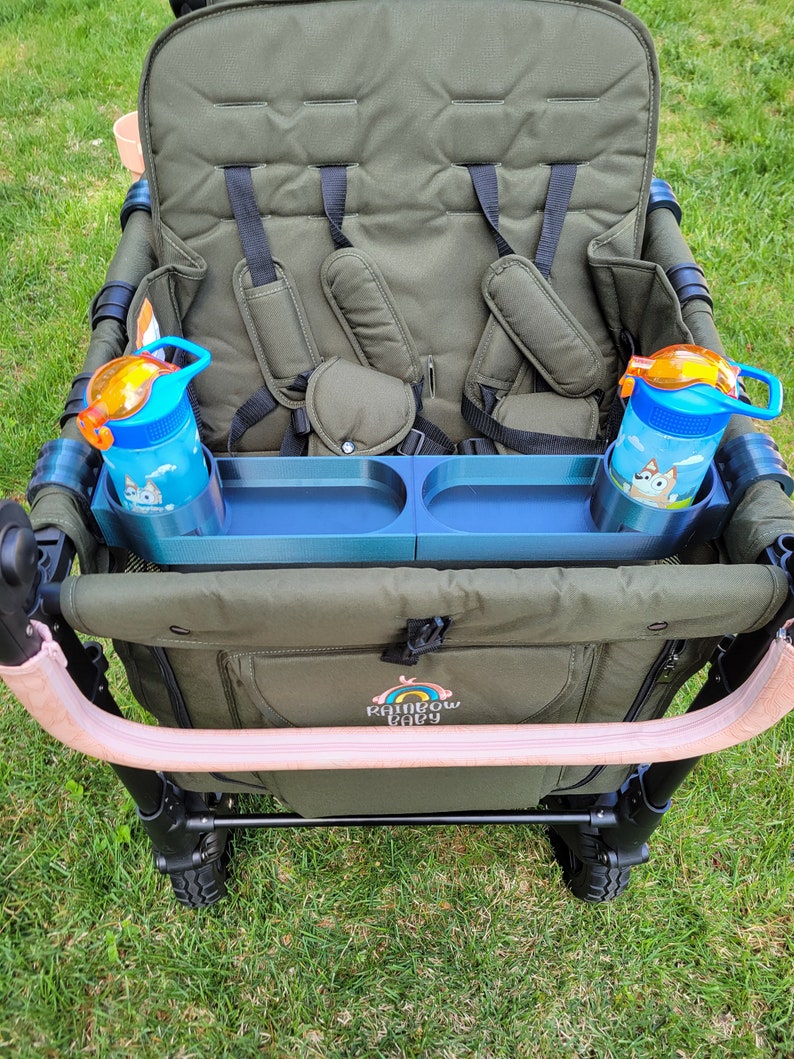 May include: A green wagon with two seats and a blue tray with two cup holders. The wagon has a pink handle and the words "Rainbow Baby" are printed on the side. Two blue water bottles with orange lids are in the cup holders.