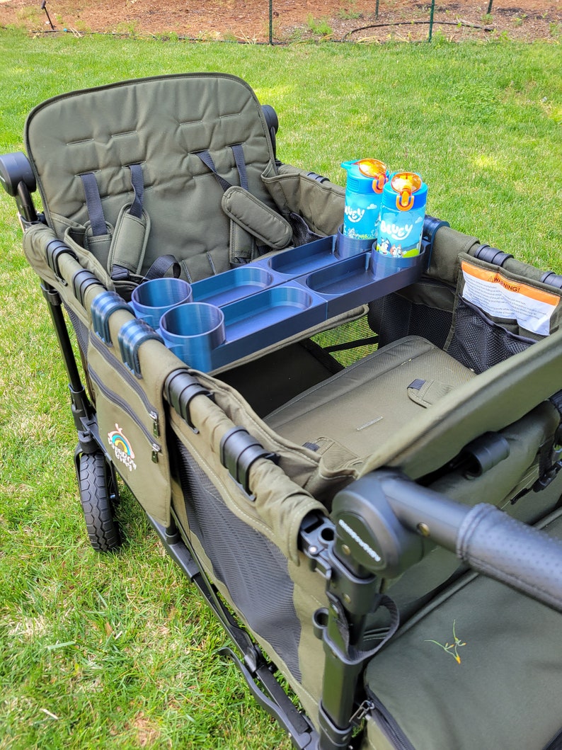 May include: A green wagon with a black frame and a black handle. The wagon has a seat with two sets of straps and a cup holder. There are two blue and orange juice boxes in the cup holder. The wagon has a black mesh pocket on the side and a black and white logo that says "Rainbow Wagon".